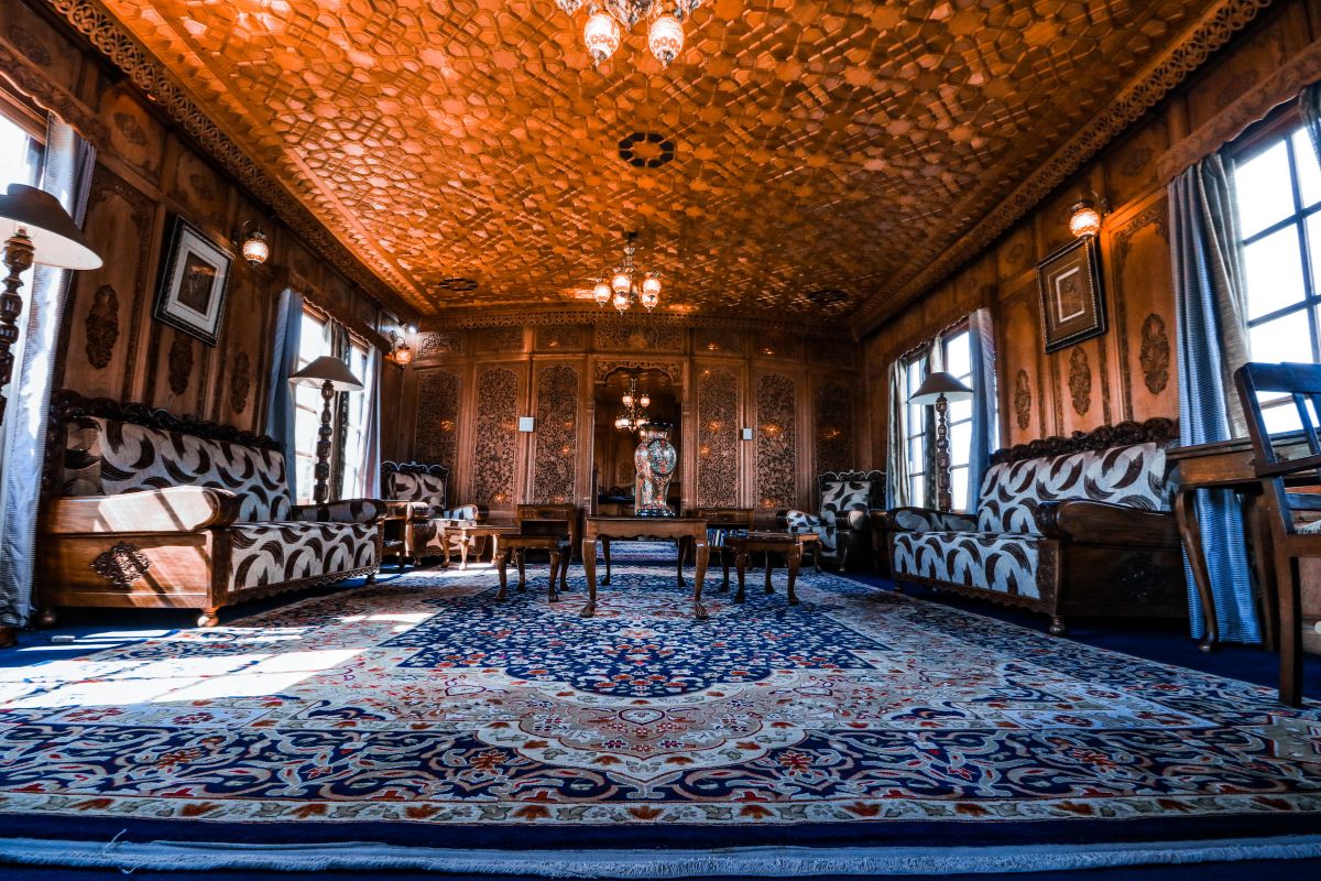 Luxurious wooden-paneled room with intricate ceiling, ornate furniture, and a patterned rug, illuminated by elegant chandeliers.