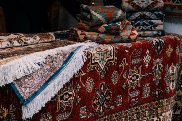 Close-up of colorful, patterned rugs and textiles stacked on a table, showcasing intricate designs and fringed edges.